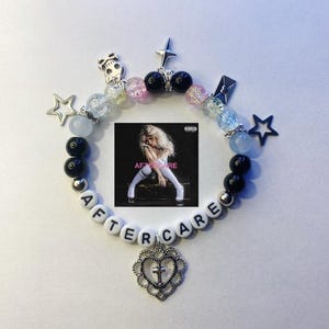 May include: A white beaded bracelet with black beads and silver charms, including stars, a skull, a heart with a cross, and a letter 'e'. The bracelet spells out "AFTER CARE" and features a small album cover with the text "AFTERCARE" in pink.