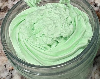 May include: A clear glass jar filled with light green, whipped cream-like substance. The cream is swirled in a decorative pattern, with a slightly darker shade of green in the center. The jar sits on a speckled surface.
