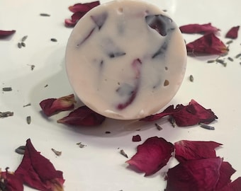 Puede incluir: Una pastilla de jabón redonda blanca con pétalos de rosa rojos incrustados. El jabón está sobre una superficie blanca con pétalos de rosa rojos esparcidos alrededor.