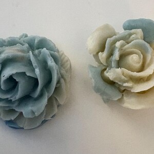 Pode incluir: Dois itens decorativos em forma de rosa. Um é principalmente azul claro, o outro é branco sujo com detalhes em azul claro. As pétalas são detalhadas e o design geral é floral. Os itens são provavelmente para artesanato ou decoração.