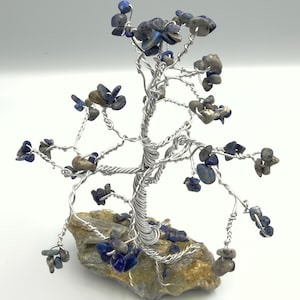May include: A decorative wire tree sculpture featuring twisted silver-colored metal branches adorned with small, tumbled blue and grey stone chips. The tree is anchored to a rough, natural-looking stone base, with additional stone chips scattered at the base.