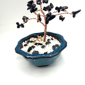 May include: A decorative bonsai tree sculpture. The tree features a copper wire trunk and branches, adorned with black gemstone leaves. It is planted in a blue ceramic pot filled with white pebbles and black stones.