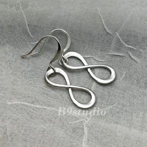 May include: A pair of silver-colored infinity symbol earrings. The earrings are displayed on a textured, light gray fabric. The infinity symbols are smooth and have a polished finish. The hooks are curved.