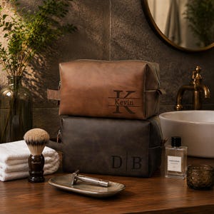 May include: Two rectangular leather wash bags stacked on a wooden surface. The top bag is brown with the name Kevin and a large letter K engraved. The bottom bag is dark grey with the initials D B engraved. Both feature side handles.