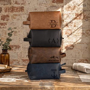 Puede incluir: Una pila de cuatro neceseres rectangulares en color tostado, negro, marr&oacute;n y azul marino. Cada bolsa cuenta con un asa lateral y un grabado personalizado &uacute;nico: 'B Burton', 'A ADAM' con laureles, 'Mike Wilson' con monta&ntilde;as y 'DANIEL' con un ancla.