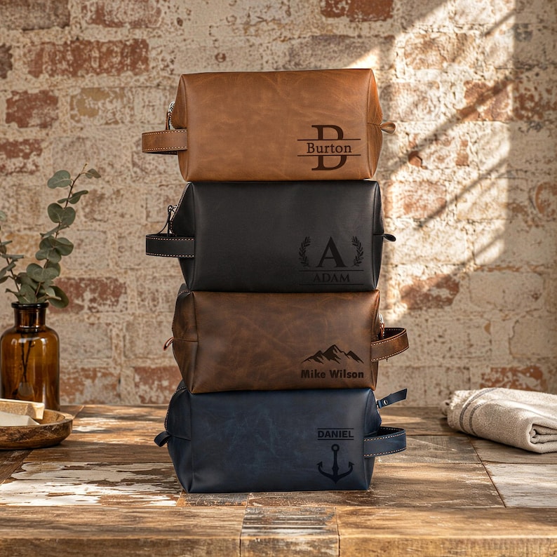 Puede incluir: Una pila de cuatro neceseres rectangulares en color tostado, negro, marr&oacute;n y azul marino. Cada bolsa cuenta con un asa lateral y un grabado personalizado &uacute;nico: 'B Burton', 'A ADAM' con laureles, 'Mike Wilson' con monta&ntilde;as y 'DANIEL' con un ancla.