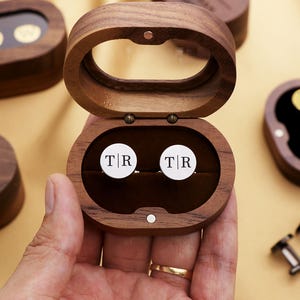 May include: Wooden oval-shaped cufflink box with a hinged lid, open to reveal two white cufflinks with the initials "TR". The box is made of dark wood and has a dark brown velvet interior. Other boxes and cufflinks are visible in the background.