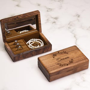 Peut inclure: Une boîte à bijoux en bois avec un couvercle à charnière et un miroir à l'intérieur. La boîte est ouverte, révélant des bagues et un collier de perles. La boîte fermée porte le nom "Diana" gravé sur le couvercle, avec des accents de feuilles.