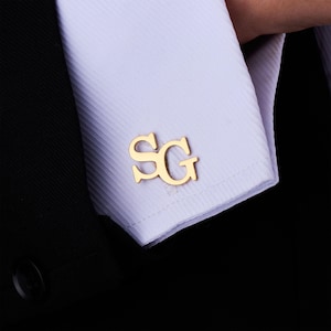 May include: Gold-colored cufflinks with the initials "SG" displayed on a white dress shirt cuff. The cufflinks are in focus, with the shirt and a black suit jacket in the background. The cufflinks are a classic accessory.