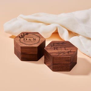 May include: Two hexagonal wooden ring boxes with engraved details. One box features "D & S" with a date and names, the other has "R L" and a date. Both boxes are brown with a dark wood grain and a decorative leaf design.