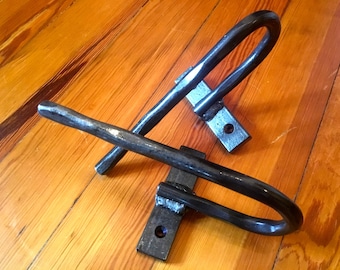 May include: Two black metal towel hooks with a curved design and a rectangular base. The hooks are mounted on a wooden surface.