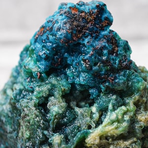 May include: A close-up of a mineral specimen with a textured surface. The mineral features a gradient of colors, transitioning from a deep teal at the top to a lighter green at the base. Small patches of brown are visible.