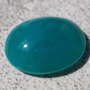 May include: A smooth, oval-shaped, teal-colored gemstone. The polished surface reflects light, creating highlights. The stone is set against a textured, light-colored background.