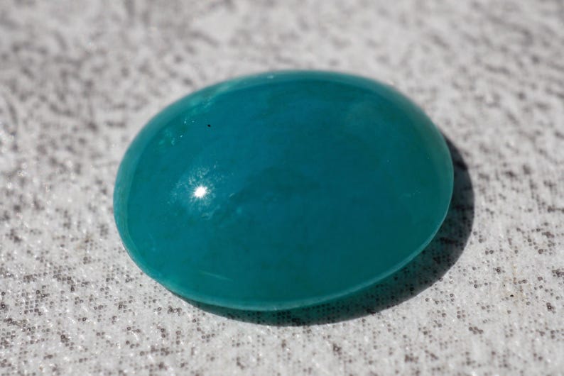 May include: A smooth, oval-shaped, teal-colored gemstone. The polished surface reflects light, creating highlights. The stone is set against a textured, light-colored background.