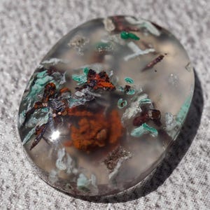 May include: A clear, oval-shaped agate cabochon with inclusions of green, brown, and white minerals. The stone has a translucent appearance and is set against a gray background.