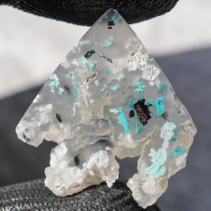 Pode incluir: Um pingente de pedra preciosa translúcida em forma triangular com um padrão único. A pedra apresenta inclusões brancas, turquesas e pretas, criando um contraste marcante. O pingente é segurado contra um fundo escuro.