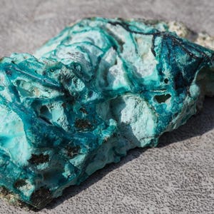 May include: A close-up of a natural stone with a striking turquoise and teal color palette. The stone has a rough, textured surface with areas of lighter, almost white, coloration. The stone's unique shape and color variations make it a visually interesting specimen.