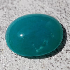 May include: A smooth, oval-shaped, teal-colored gemstone. The polished surface reflects light, creating highlights. The stone is set against a textured, light-colored background.