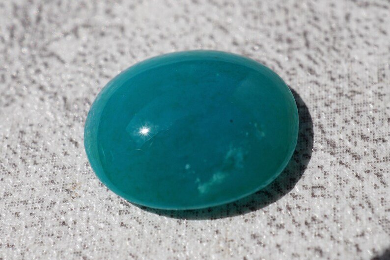 May include: A smooth, oval-shaped, teal-colored gemstone. The polished surface reflects light, creating highlights. The stone is set against a textured, light-colored background.