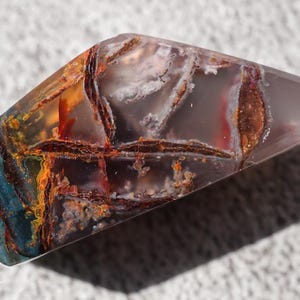 May include: A polished gemstone with a unique pattern of colors. The stone features a mix of blue, orange, brown, and white hues. The gemstone is cut into a rectangular shape with angled edges, and is likely used for jewelry or display.