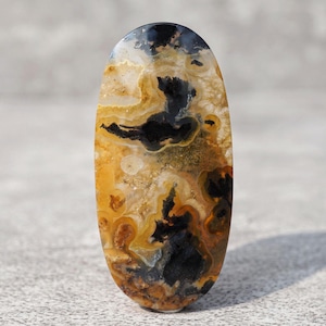 May include: An oval-shaped polished gemstone cabochon featuring a complex pattern of translucent amber, golden yellow, and opaque black inclusions. The stone displays swirling organic bands and distinct dark mineral formations throughout its surface.