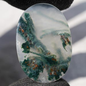 May include: An oval-shaped moss agate gemstone with a translucent, milky white base. The stone features prominent, deep green, fern-like dendritic inclusions and small, scattered copper-colored mineral deposits throughout the surface.
