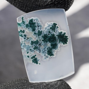 May include: A rectangular gemstone with a light gray base and a striking pattern of dark green and light blue moss-like inclusions. The stone is held against a dark, textured surface, showcasing its natural beauty.