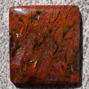 May include: A square-shaped stone with a polished surface. The stone features a rich color palette of deep reds, browns, and hints of green, with a textured pattern of diagonal lines. The stone's unique appearance suggests it could be used for jewelry or decorative purposes.
