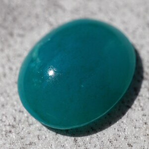 May include: A smooth, oval-shaped teal gemstone with a glossy finish. The polished surface reflects light, creating highlights. The gemstone is a vibrant shade of teal, with subtle variations in color. This gemstone could be used in jewelry.