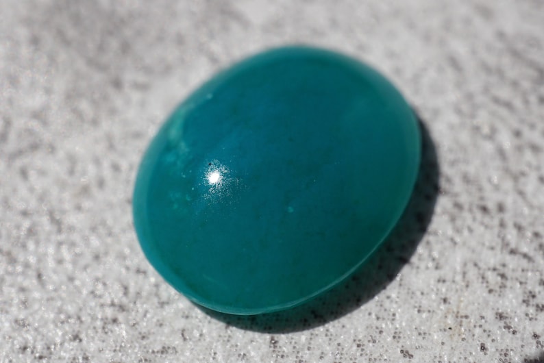 May include: A smooth, oval-shaped teal gemstone with a glossy finish. The polished surface reflects light, creating highlights. The gemstone is a vibrant shade of teal, with subtle variations in color. This gemstone could be used in jewelry.
