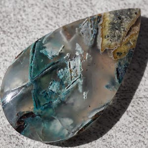 May include: A teardrop-shaped gemstone with a polished surface. The stone features a mix of colors, including teal, brown, and yellow, with a translucent, glassy appearance. The stone's unique patterns and colors make it a striking piece.