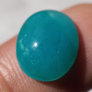 May include: A smooth, oval-shaped, turquoise gemstone. The polished stone has a glossy surface with subtle variations in color. The gemstone is held between two fingers, with a neutral background.
