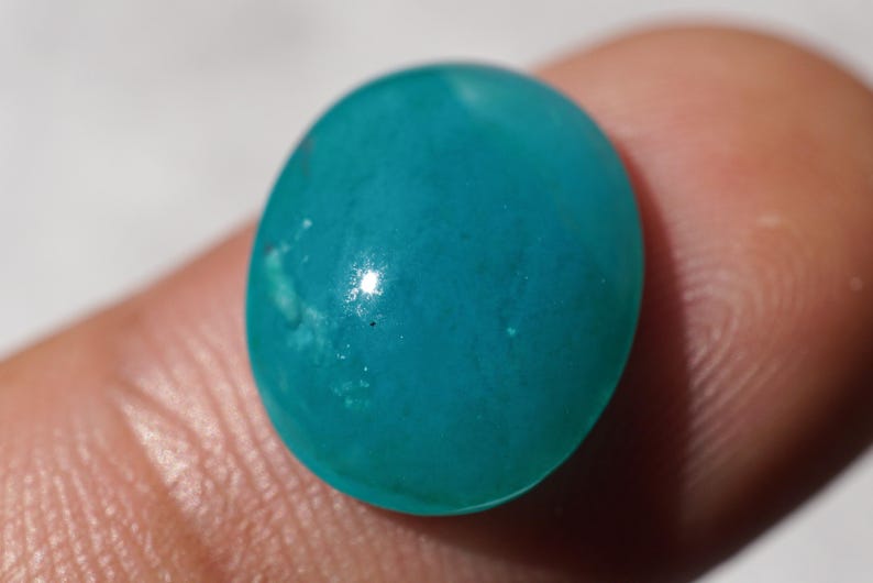 May include: A smooth, oval-shaped, turquoise gemstone. The polished stone has a glossy surface with subtle variations in color. The gemstone is held between two fingers, with a neutral background.