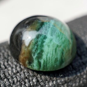 May include: A polished, dome-shaped cabochon gemstone featuring a blend of deep black, earthy brown, and vibrant teal green colors with a smooth, glossy surface that reflects light, showcasing natural swirling patterns and textures throughout.
