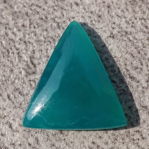 Pode incluir: Uma pedra preciosa de formato triangular com uma superfície lisa e polida de uma cor verde-azulada vibrante. A pedra apresenta uma base plana e um topo pontiagudo, com um acabamento brilhante que reflete a luz sobre o seu aspeto sólido.
