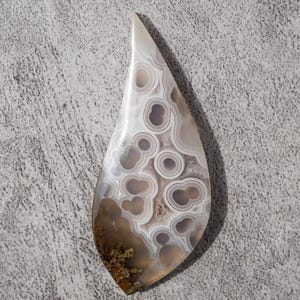May include: A teardrop-shaped polished stone with a unique pattern of concentric circles and swirls in shades of white, gray, and brown. The stone has a smooth, glossy surface and is likely a decorative item or a component for jewelry.