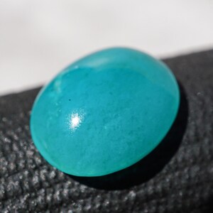 May include: A smooth, oval-shaped, turquoise gemstone. The polished surface reflects light, creating a bright spot. The gem is set against a dark, textured background, highlighting its vibrant color. This gemstone could be used in jewelry.