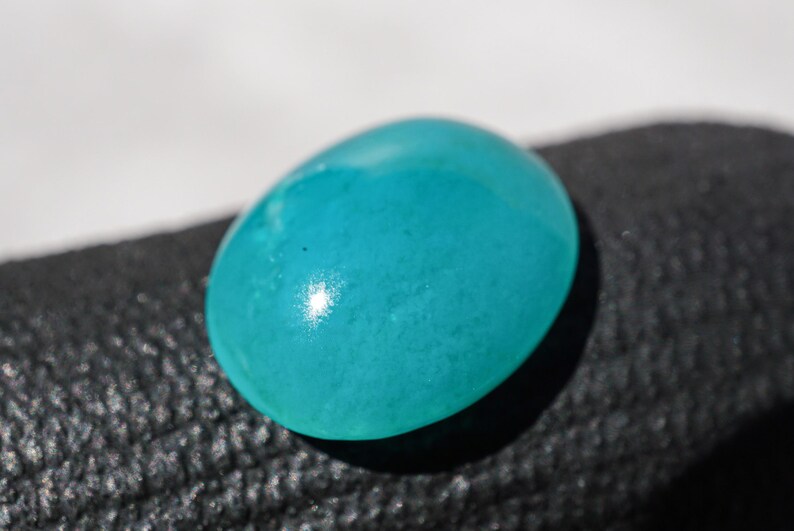 May include: A smooth, oval-shaped, turquoise gemstone. The polished surface reflects light, creating a bright spot. The gem is set against a dark, textured background, highlighting its vibrant color. This gemstone could be used in jewelry.