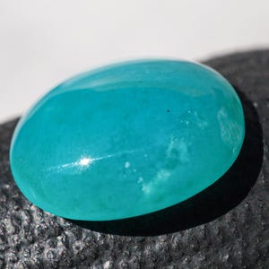 May include: A polished, oval-shaped gemstone in a vibrant turquoise hue. The stone has a smooth, glossy surface with subtle internal patterns and a bright highlight reflecting light. The gemstone is likely used in jewelry.