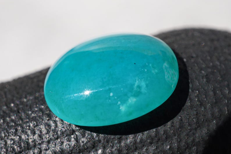 May include: A polished, oval-shaped gemstone in a vibrant turquoise hue. The stone has a smooth, glossy surface with subtle internal patterns and a bright highlight reflecting light. The gemstone is likely used in jewelry.