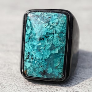 May include: A ring featuring a large, rectangular, textured turquoise stone with vibrant blue and green hues, set within a smooth, polished black metal band. The stone displays a natural, porous surface pattern with dark inclusions throughout.