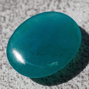 May include: A smooth, oval-shaped teal stone with a glossy finish. The stone is a vibrant shade of blue-green, and it casts a shadow on the textured surface beneath it. The gemstone is likely used for jewelry or decorative purposes.