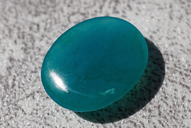 May include: A smooth, oval-shaped teal stone with a glossy finish. The stone is a vibrant shade of blue-green, and it casts a shadow on the textured surface beneath it. The gemstone is likely used for jewelry or decorative purposes.