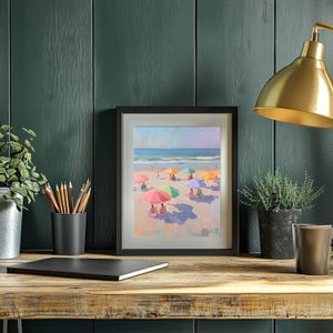 May include: A framed painting of a beach scene with colorful umbrellas and people relaxing on the sand. The painting is in a black frame and is hanging on a green wall.
