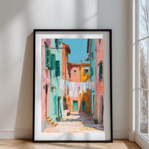 May include: A framed art print of a colorful Italian street scene. The artwork depicts buildings in shades of pink, turquoise, and orange, with laundry hanging between them. The sky is a bright blue, and the overall style is painterly.