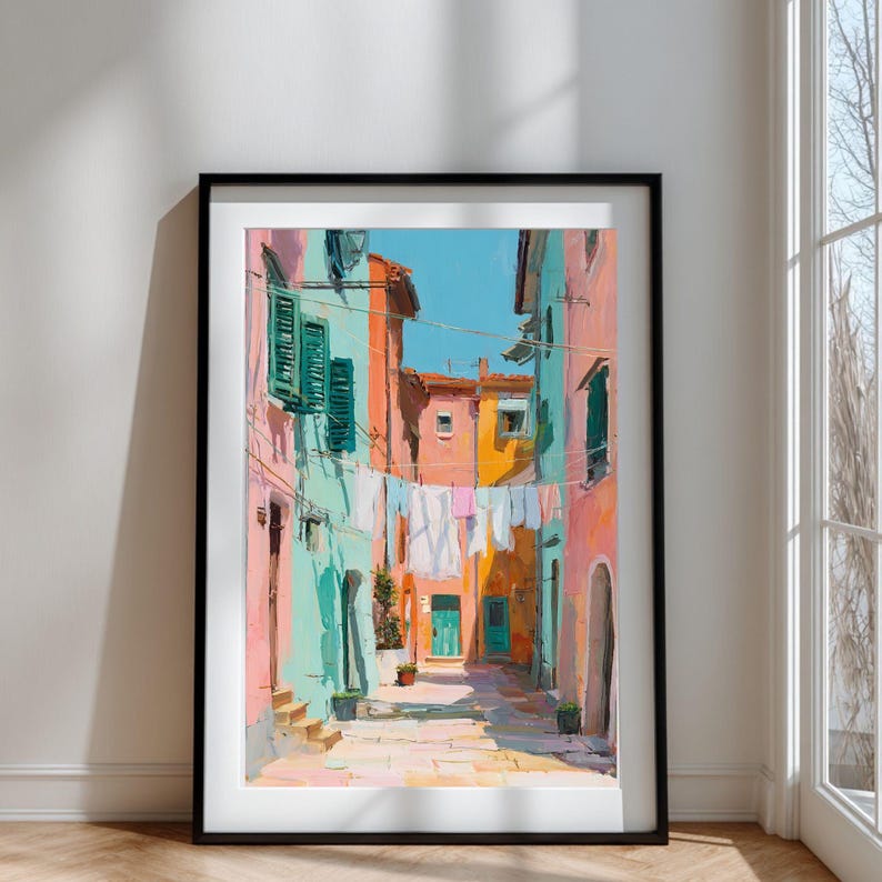 May include: A framed art print of a colorful Italian street scene. The artwork depicts buildings in shades of pink, turquoise, and orange, with laundry hanging between them. The sky is a bright blue, and the overall style is painterly.