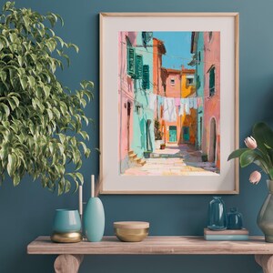 May include: A framed art print depicting a colorful Italian street scene. The artwork features buildings in shades of pink, orange, and turquoise, with laundry hanging between them. The sky is a bright blue. The print is displayed on a wooden shelf with decorative items.