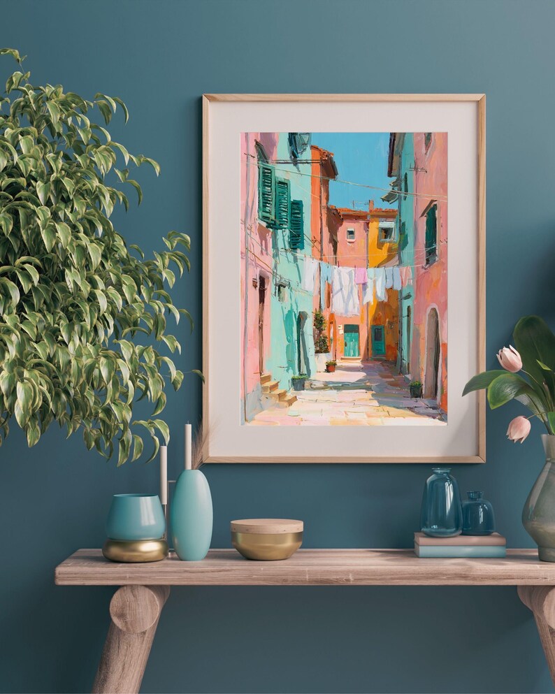 May include: A framed art print depicting a colorful Italian street scene. The artwork features buildings in shades of pink, orange, and turquoise, with laundry hanging between them. The sky is a bright blue. The print is displayed on a wooden shelf with decorative items.