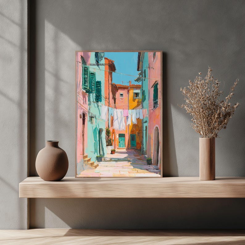 May include: A framed art print depicting a colorful Italian street scene. The artwork features buildings in shades of pink, orange, and turquoise, with laundry hanging between them. The sky is a bright blue, and the scene is bathed in sunlight.