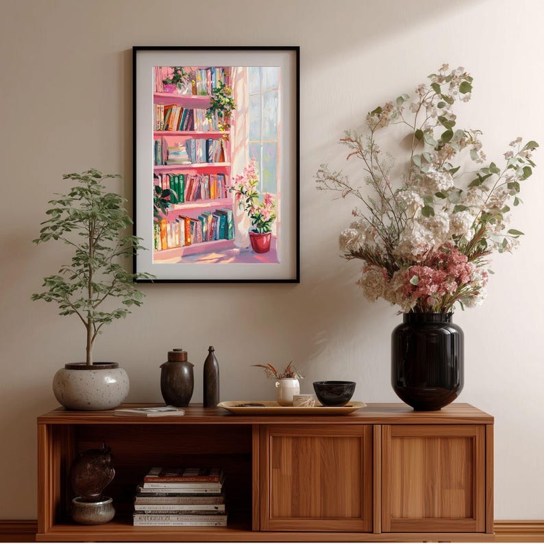 K&ouml;nnte beinhalten: Ein gerahmtes Gem&auml;lde zeigt ein rosa B&uuml;cherregal mit bunten B&uuml;chern, Pflanzen und Sonnenlicht, das durch ein Fenster scheint. Es h&auml;ngt &uuml;ber einem Holzschrank mit Dekorationen und einer Topfpflanze.